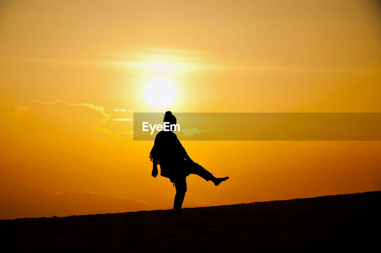 Silhouette woman standing on one leg at beach against orange sky during sunset