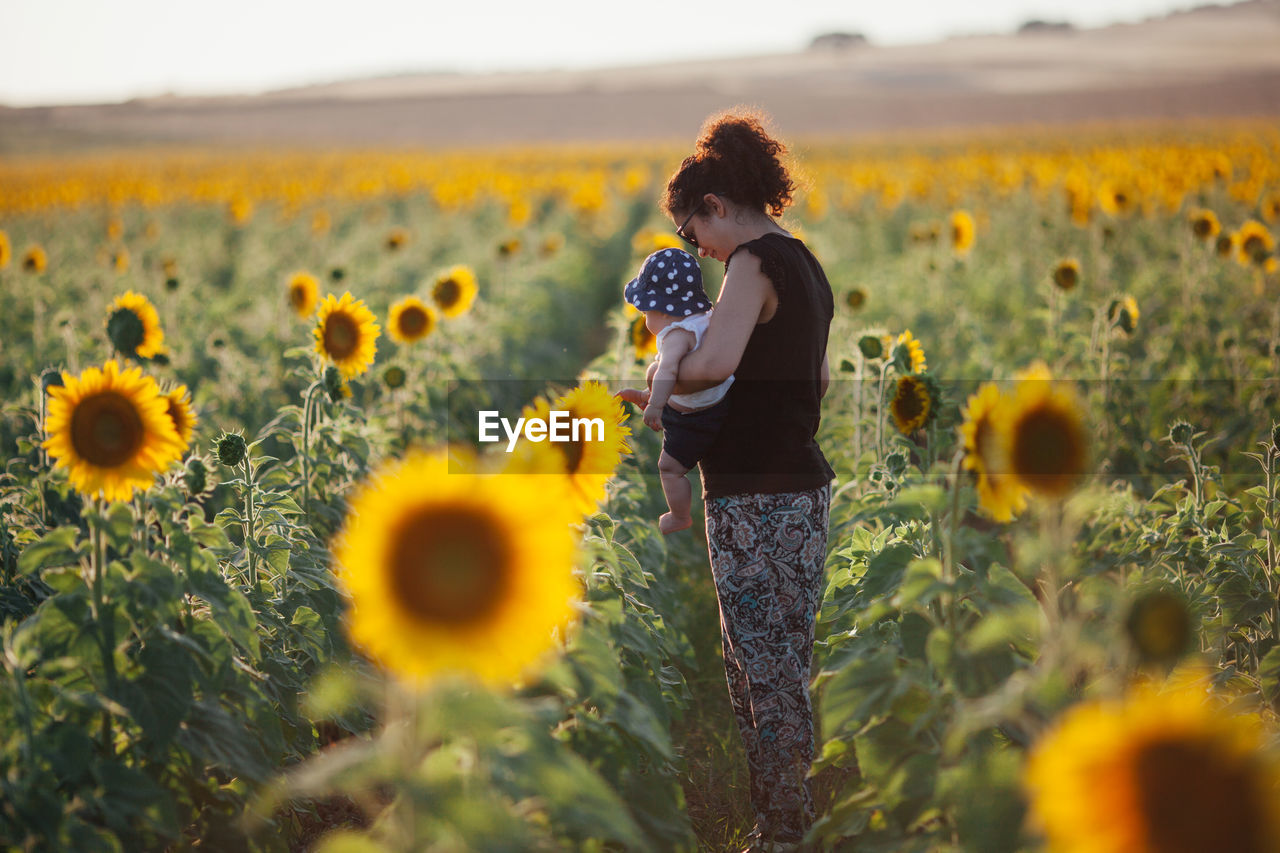 Side view of mother carrying daughter while standing amidst sunflowers in field against sky