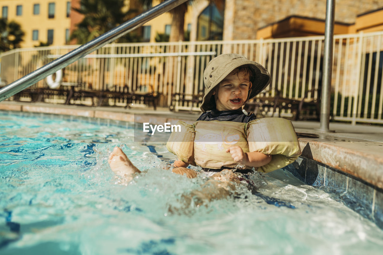 Young toddler boy splashing himself in pool and making a funny face