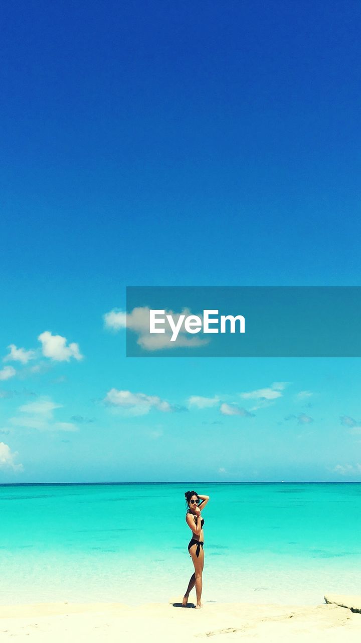 Young woman standing on shore at beach against blue sky