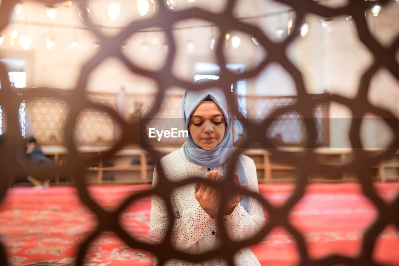 Young woman praying in mosque seen through window fence