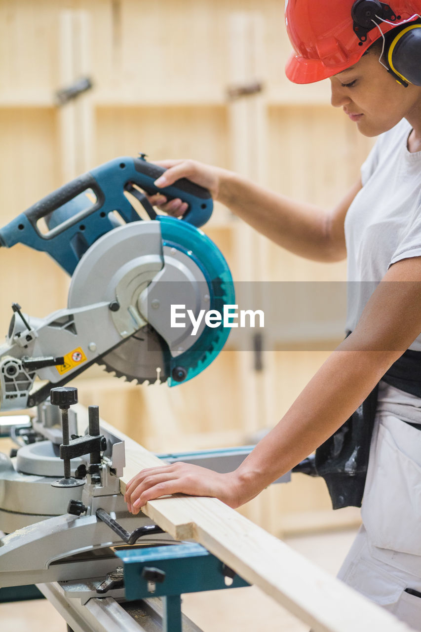 Young female trainee using electric saw for cutting plank at illuminated workshop