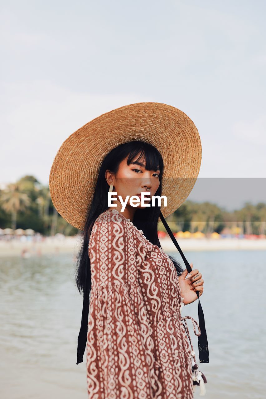 Portrait of young woman wearing hat standing at beach against sky