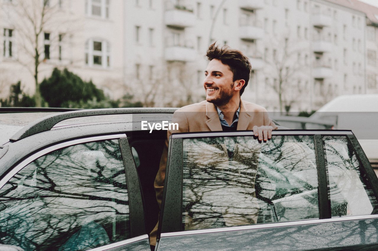 Happy young man entering wet car in city during rainy season