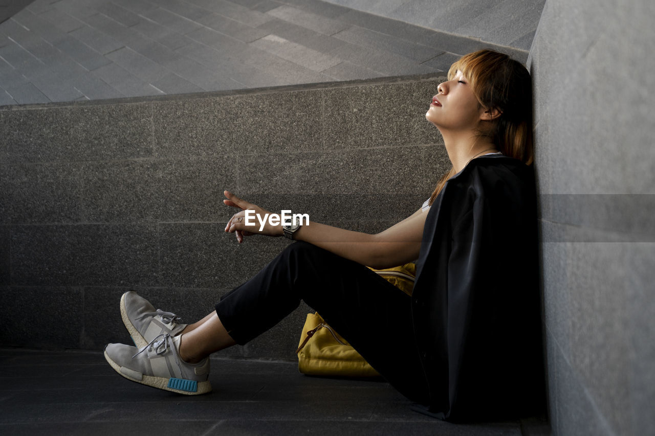 Side view of businesswoman with eyes closed sitting by wall