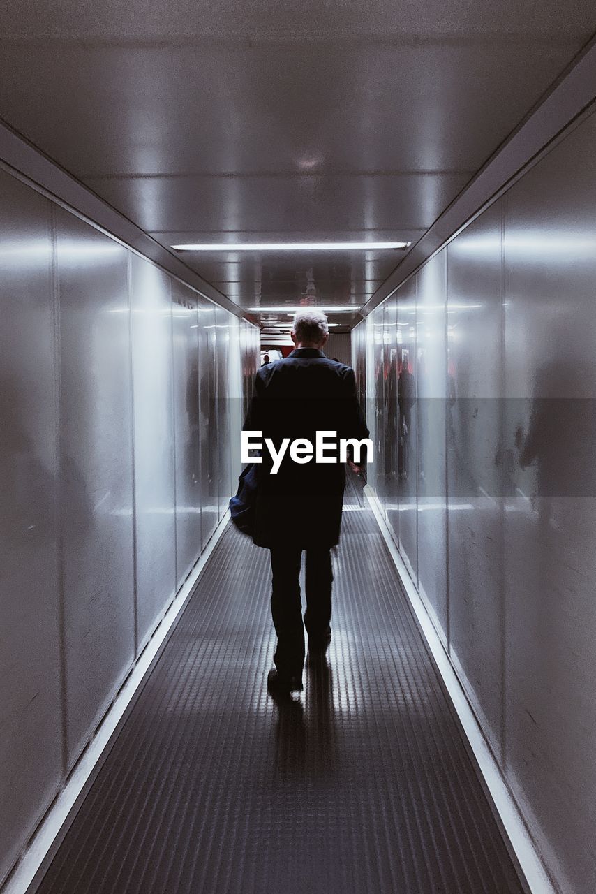 REAR VIEW OF MAN WALKING IN ILLUMINATED UNDERGROUND WALKWAY