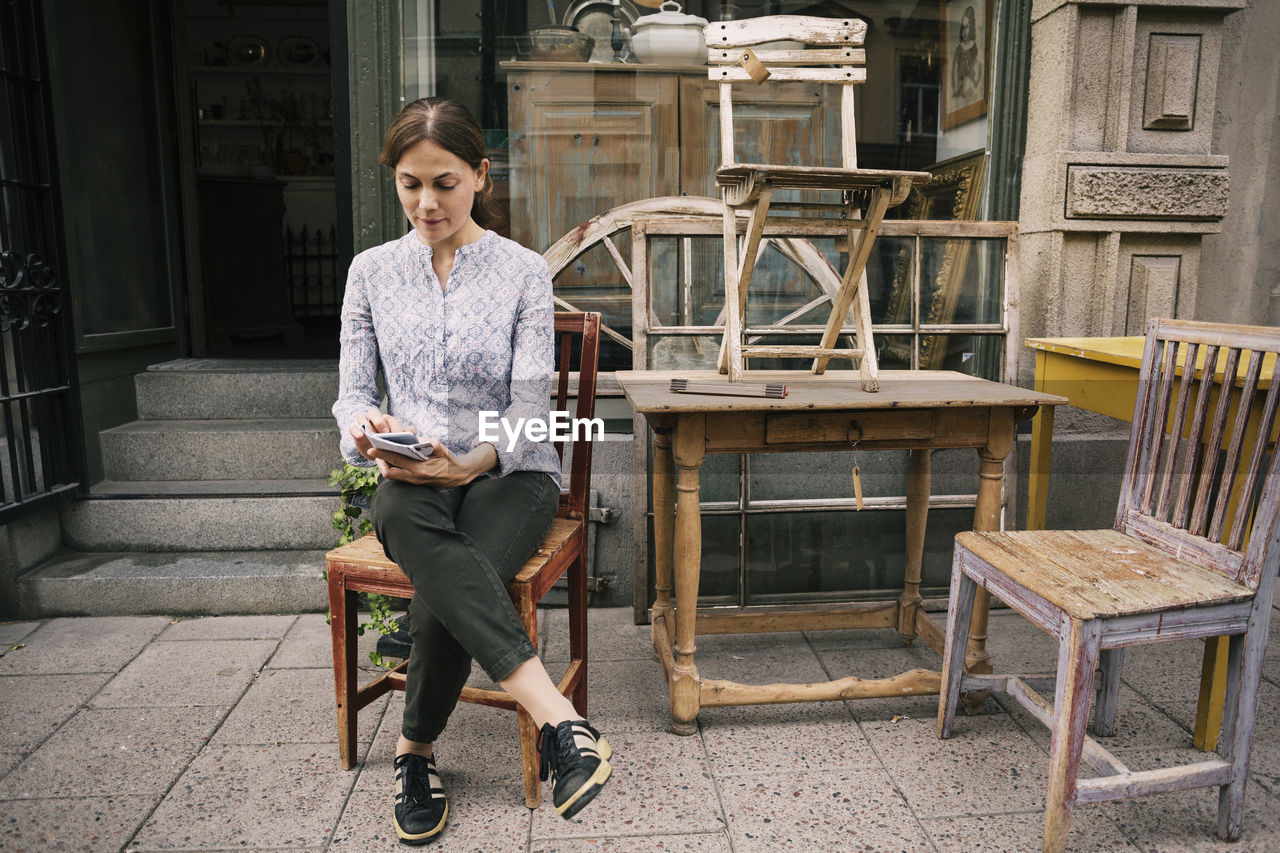 Woman using mobile phone while sitting on chair against store