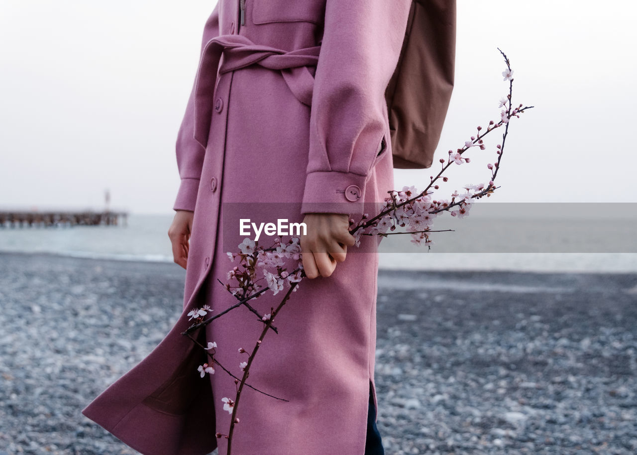 Cropped unrecognizable female in pink coat standing with blooming cheery flower twig