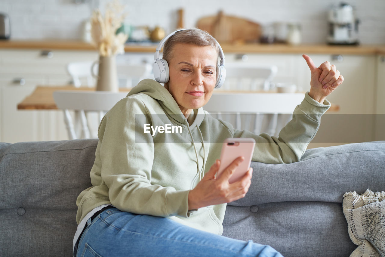 Happy senior woman in headphones listening music on smartphone while relaxing at home on sofa