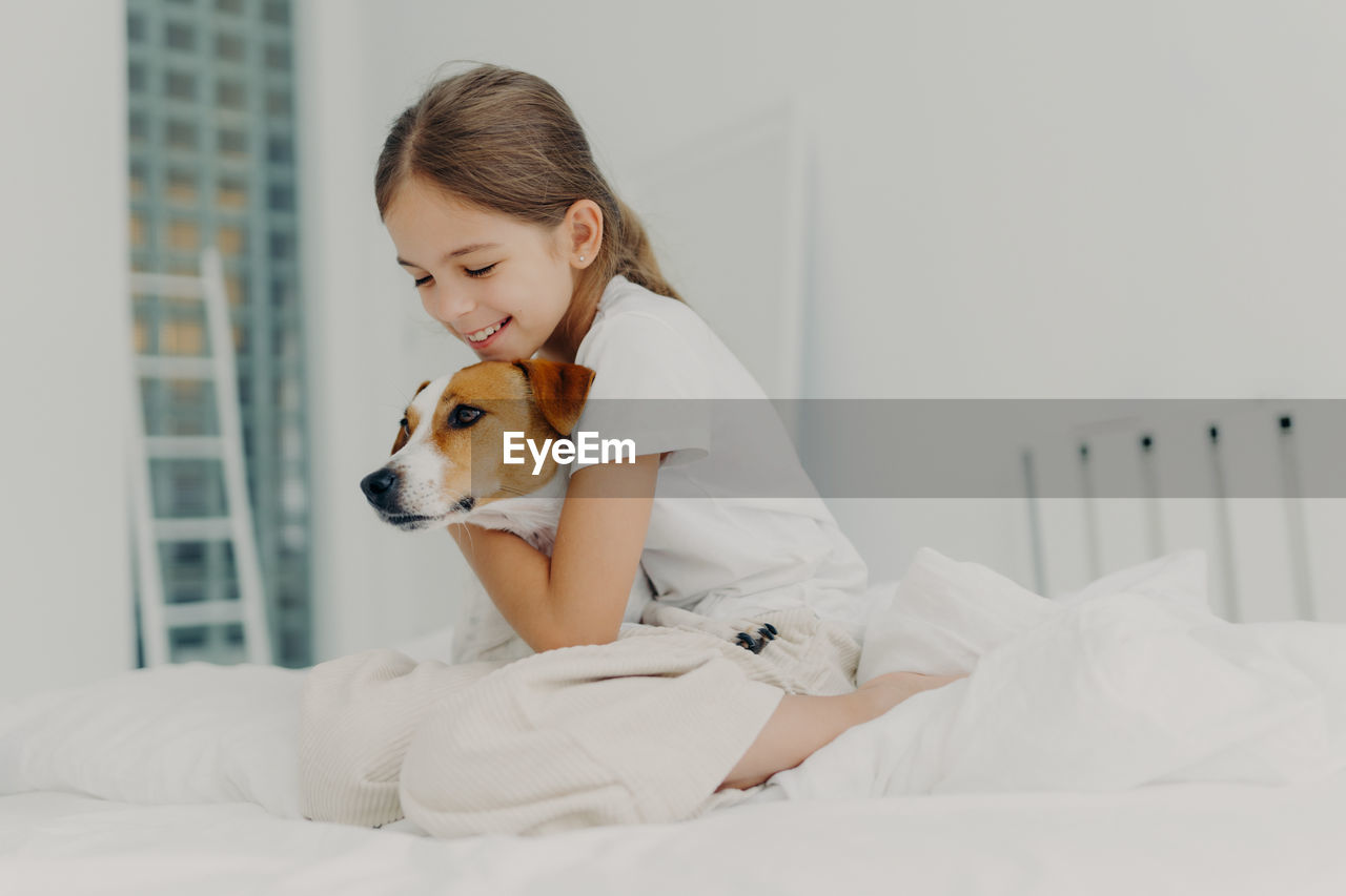 Cute girl embracing dog while sitting on bed at home