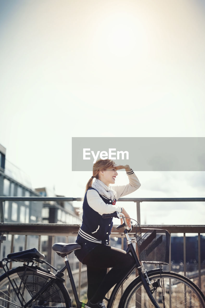Happy businesswoman shielding eyes while standing with bicycle on bridge against sky