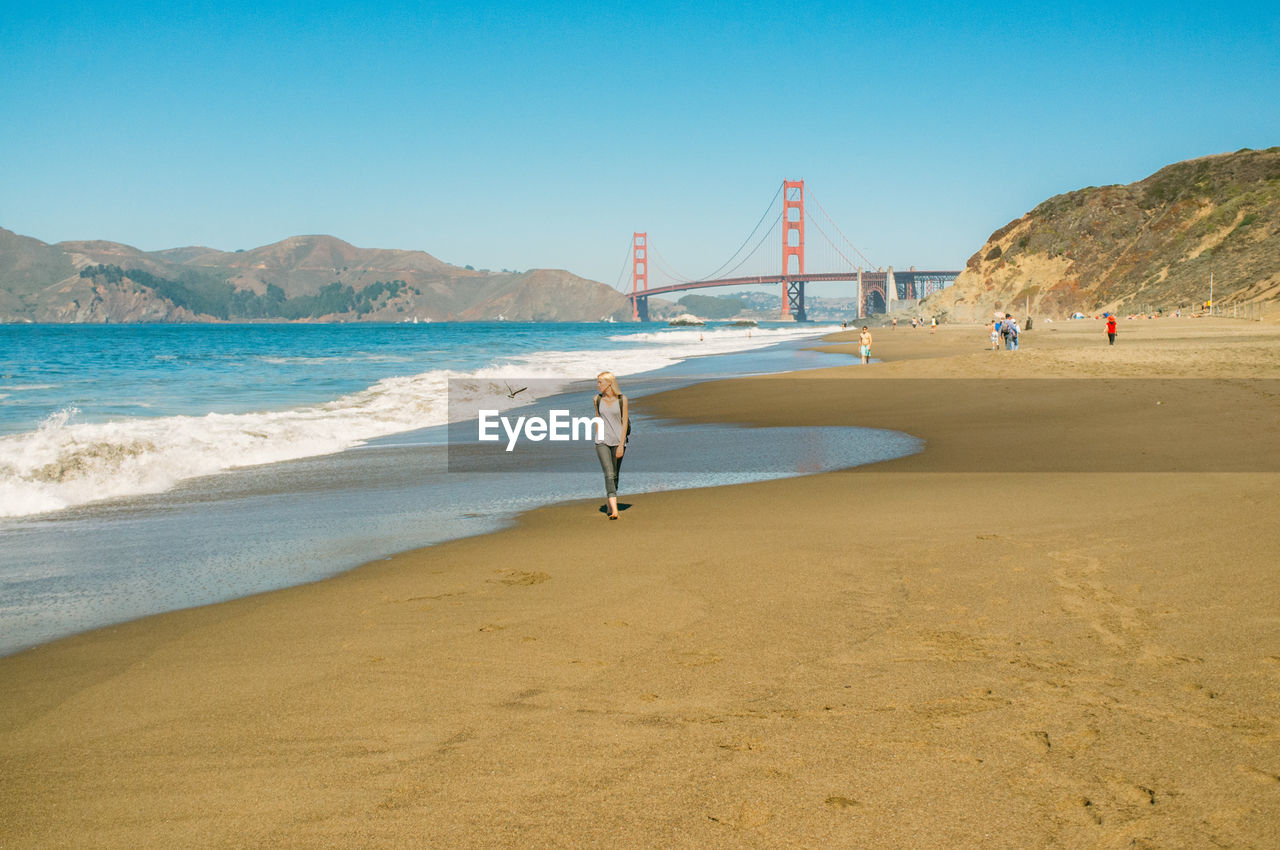 Woman walking on shore with golden gate bridge against clear sky