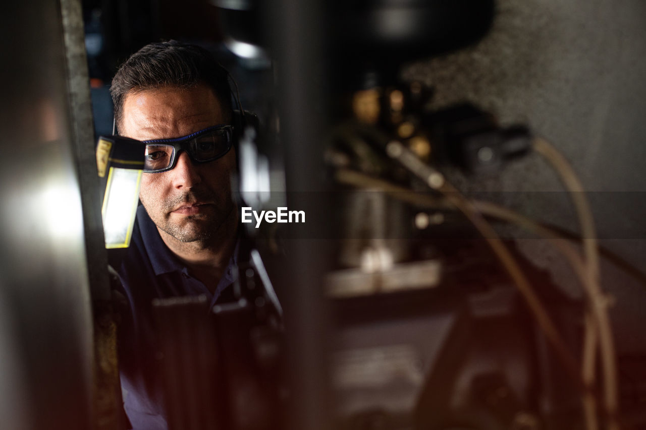 Professional male mechanic in protective glasses and headphones using flashlight while checking details of machine during repair works in workshop