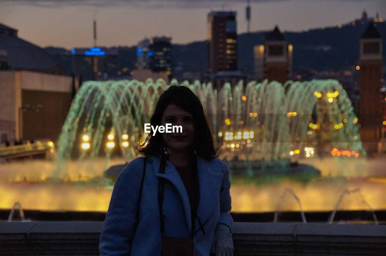 Portrait of smiling young woman standing against illuminated fountain at night