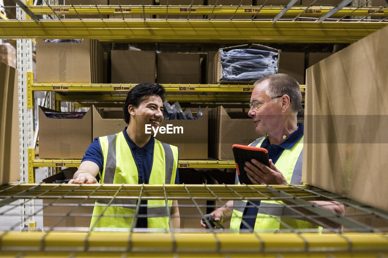 Smiling male coworkers discussing while seen through rack at warehouse