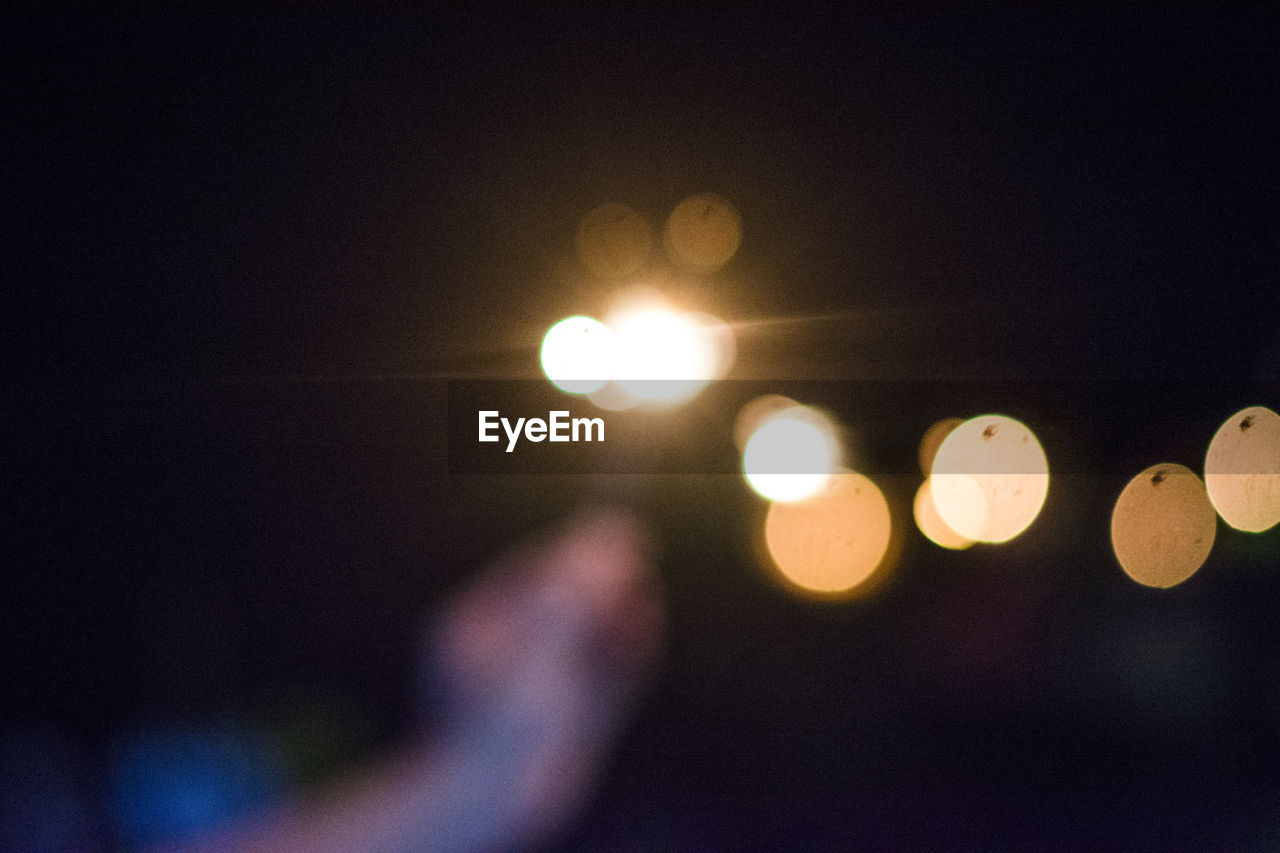 DEFOCUSED IMAGE OF HAND AGAINST ILLUMINATED LIGHTS AT NIGHT