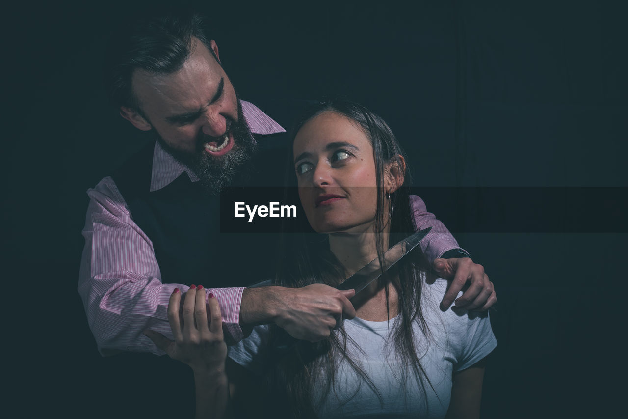 Angry man shouting while holding knife by woman against black background