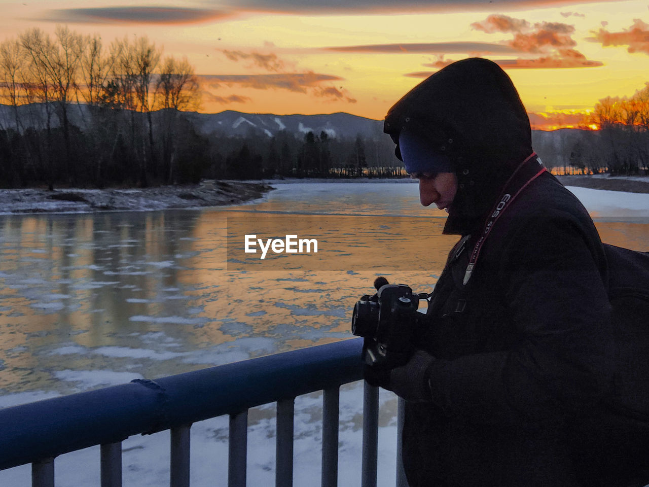 SIDE VIEW OF MAN PHOTOGRAPHING BY RAILING DURING SUNSET