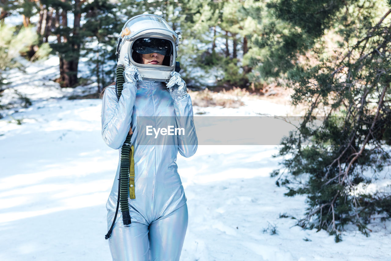 Focused unrecognizable young female astronaut in spacesuit and helmet standing in snowy woodland