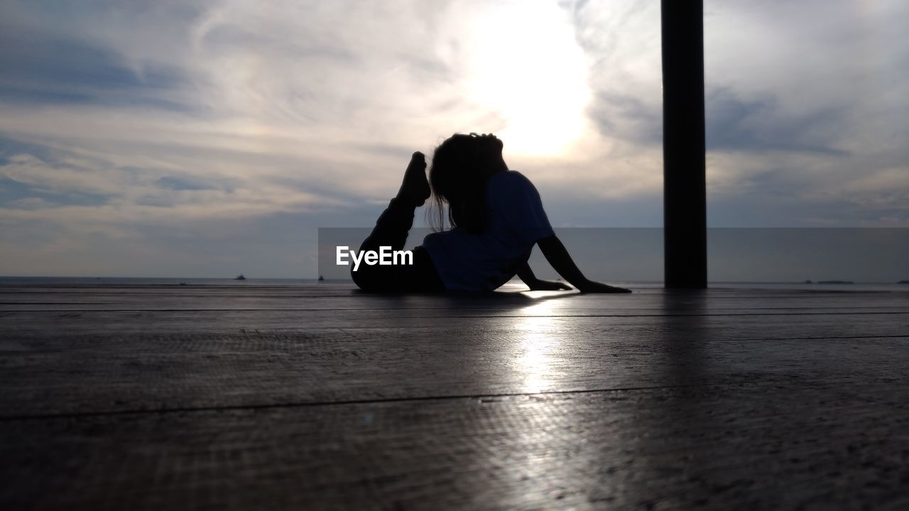 Side view of girl practicing yoga on wooden floor against cloudy sky