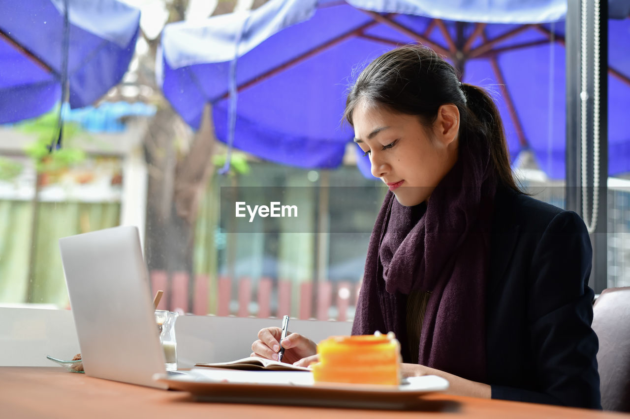 Young woman writing in book by laptop on table