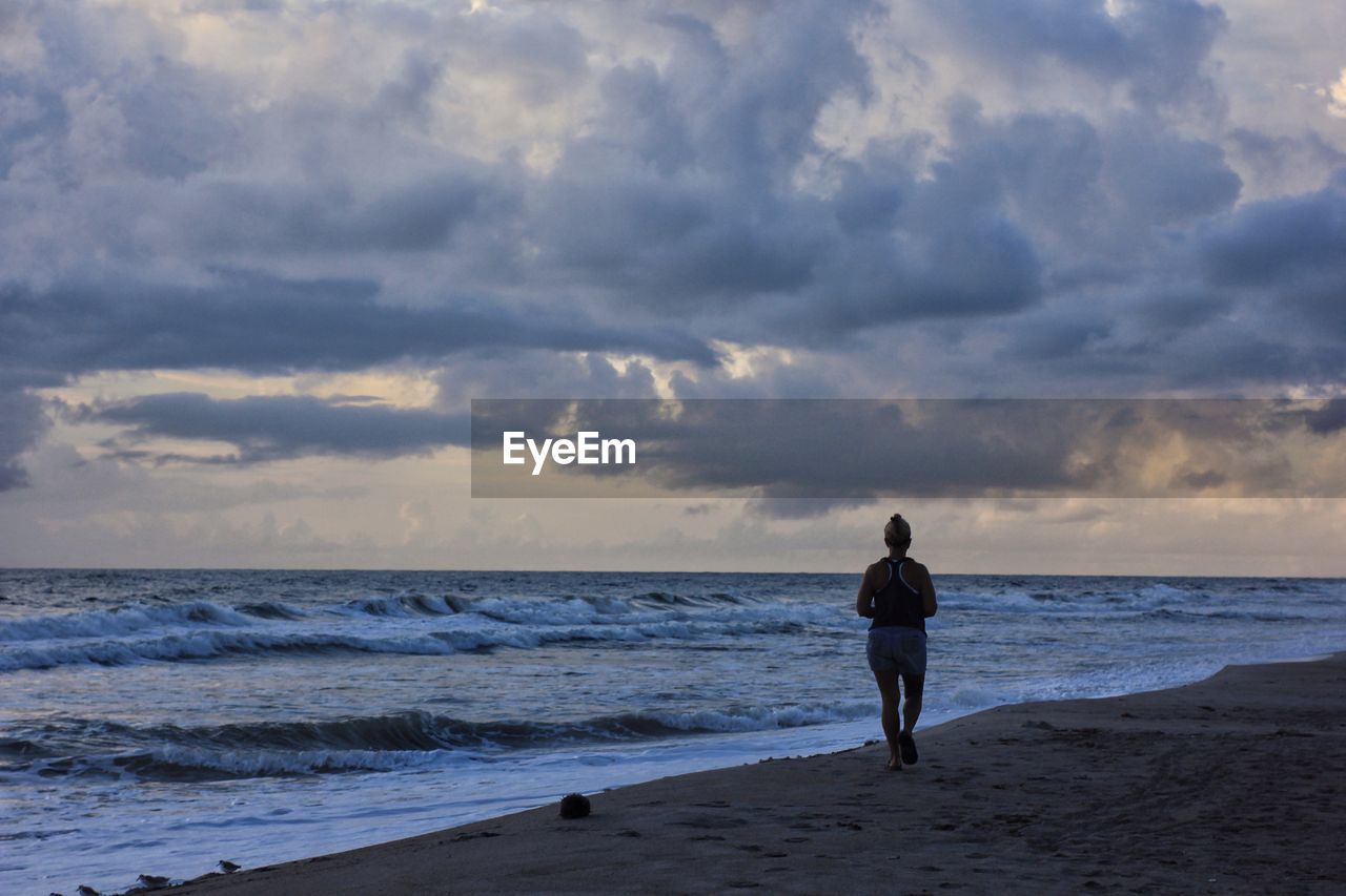 Rear view of woman walking at beach against cloudy sky during sunset