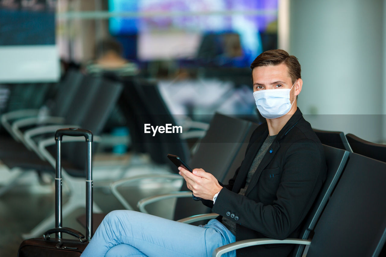 Portrait of man wearing mask using mobile sitting at airport