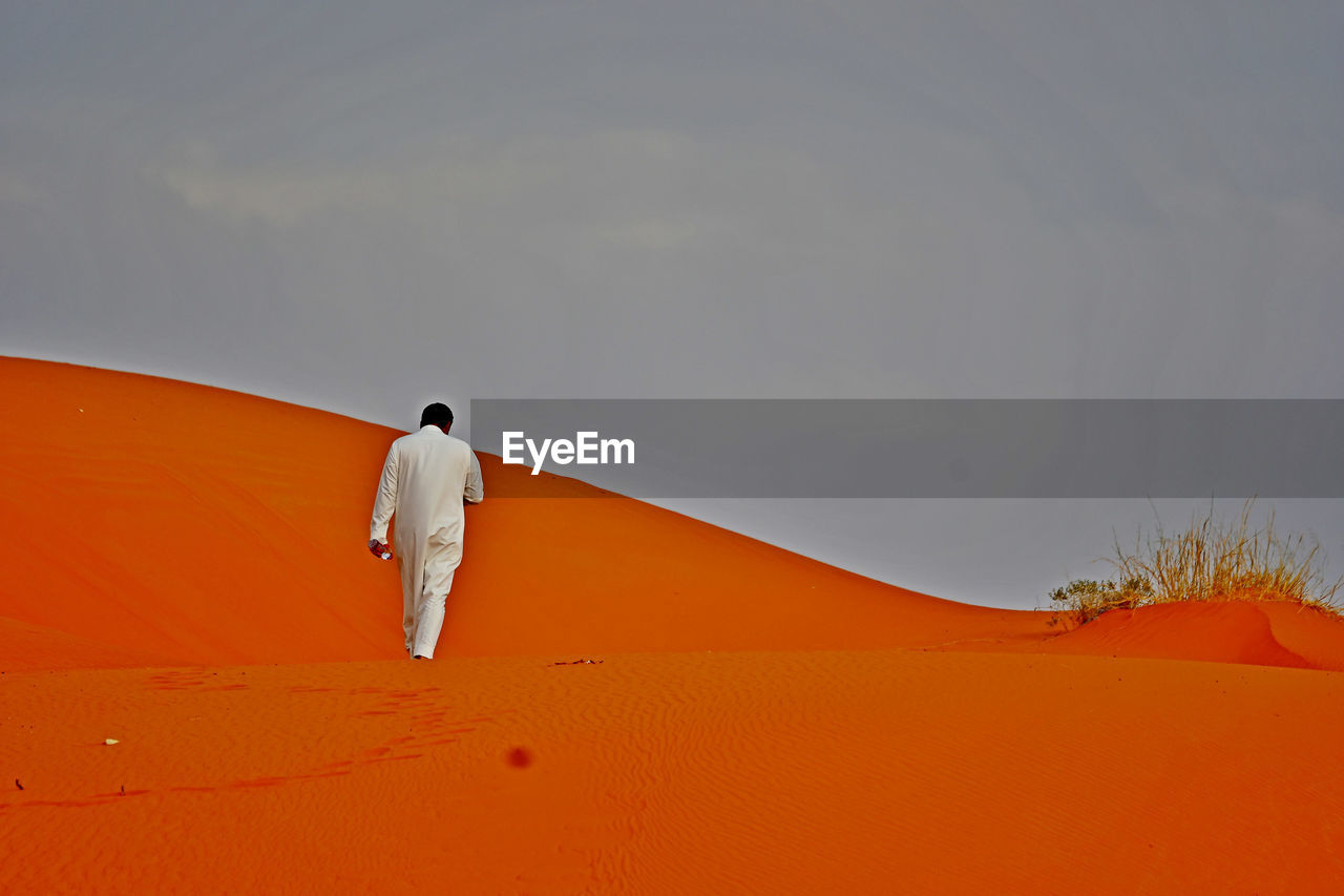 FULL LENGTH REAR VIEW OF MAN WALKING ON SAND DUNE