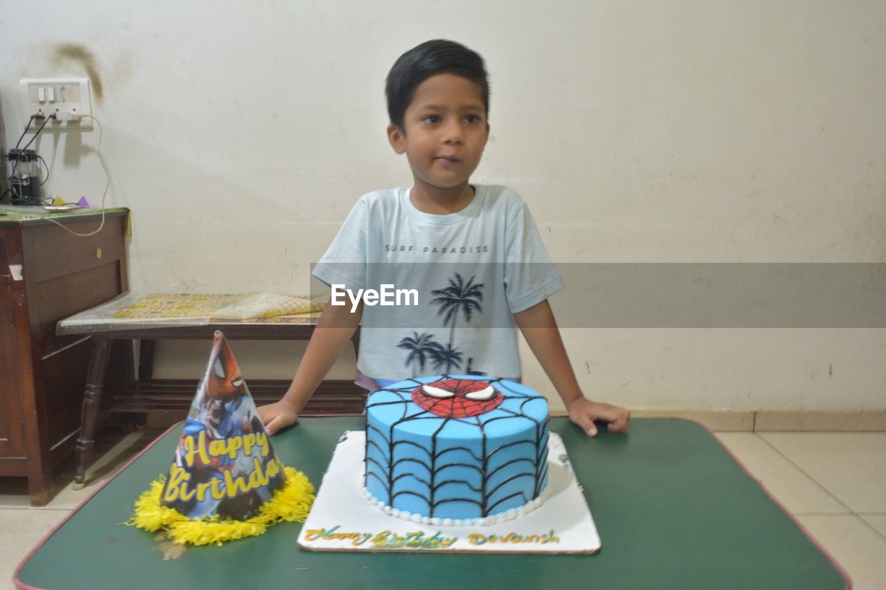 one person, child, childhood, looking at camera, portrait, front view, birthday cake, men, indoors, person, standing, smiling, emotion, table, casual clothing, lifestyles, art, innocence, toddler, sitting, cake, happiness, waist up, cute, clothing, three quarter length