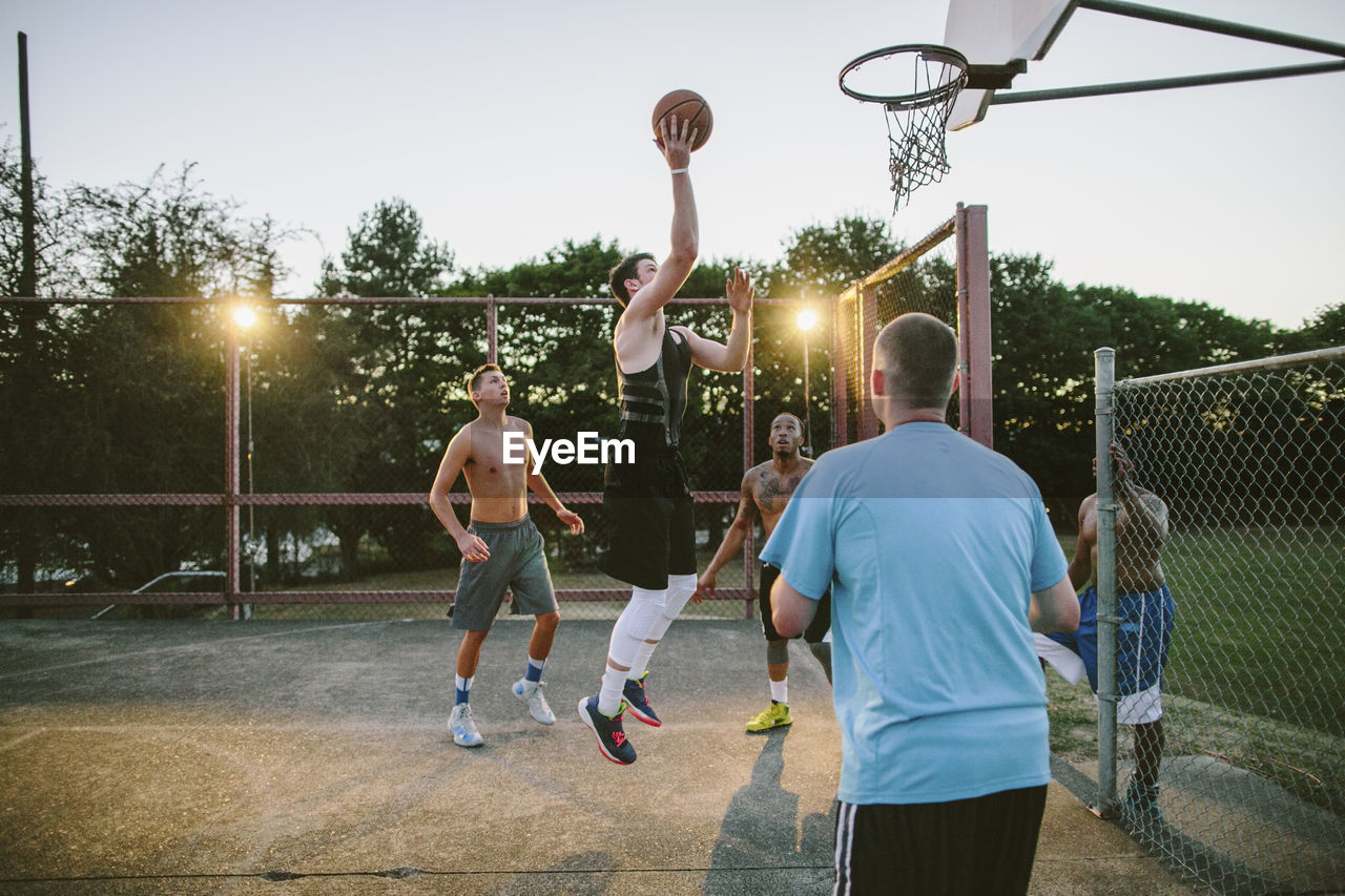 Friends looking at man dunking while practicing basketball in court during sunset