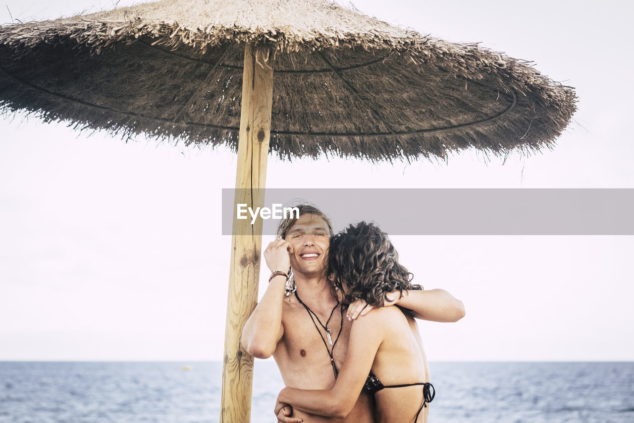 Portrait of smiling young man standing with woman by thatched roof parasol at beach