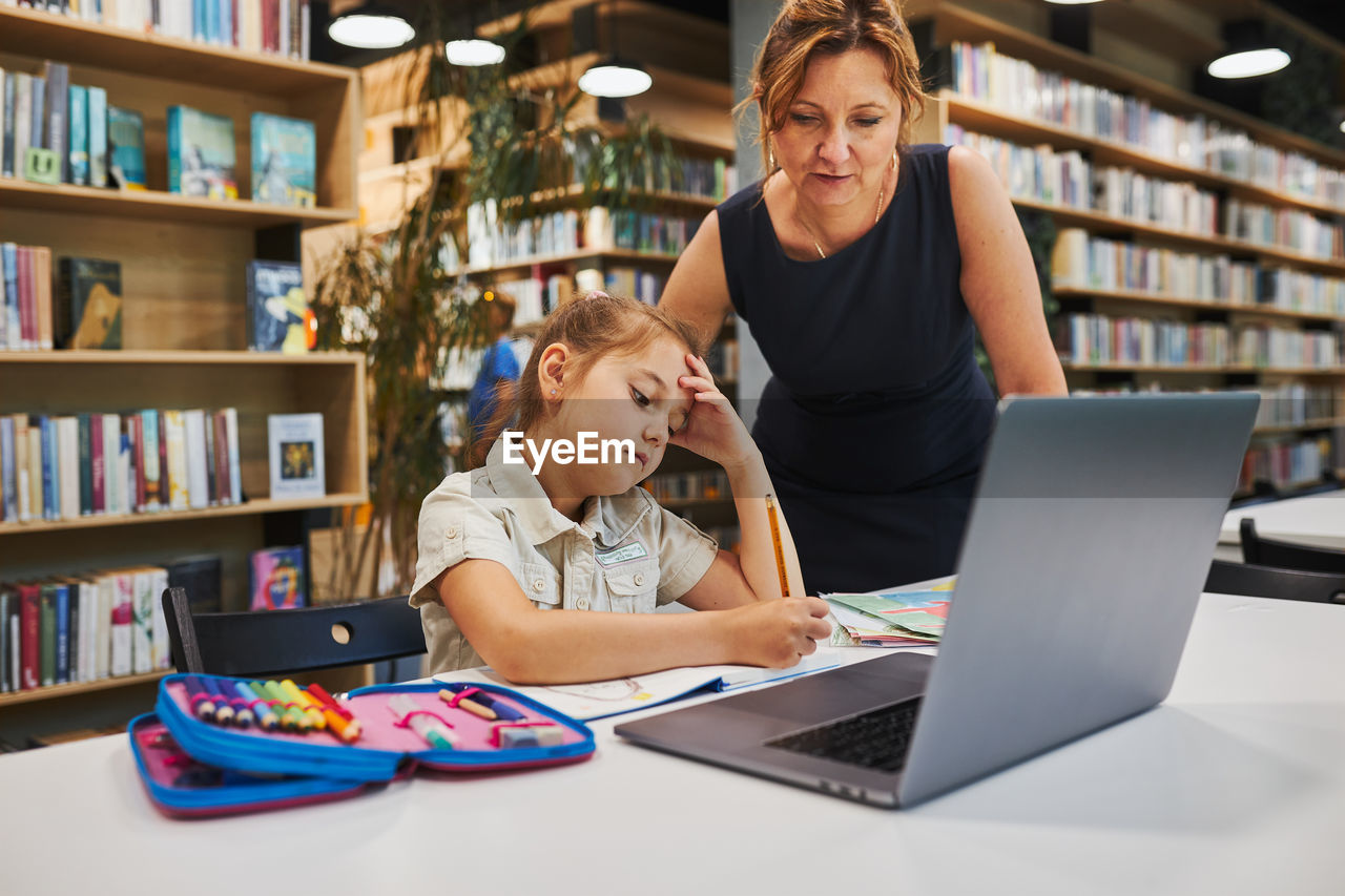 Teacher helping to solve and finish task. bored little student girl doing her homework using laptop