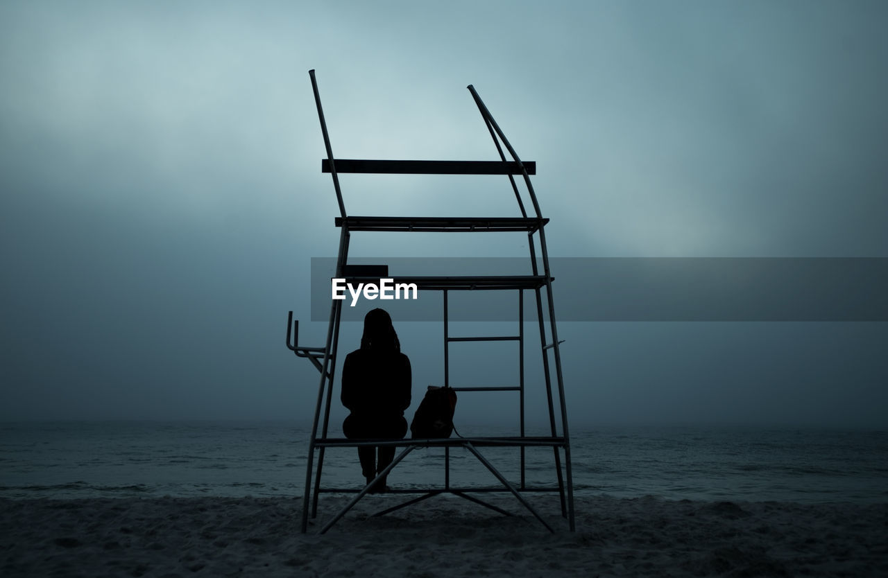 Rear view of silhouette woman sitting on built structure at beach