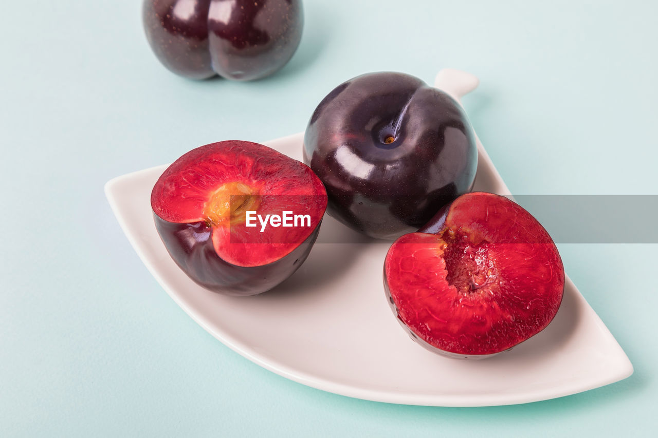 On a white saucer is a large plum, cut in half, and next to it is a whole plum.