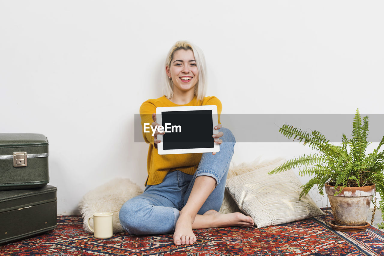 one person, technology, smiling, women, adult, sitting, indoors, wireless technology, blond hair, happiness, communication, furniture, person, computer, emotion, domestic room, lifestyles, portrait, clothing, casual clothing, front view, digital tablet, houseplant, young adult, living room, home interior, looking at camera, internet, cheerful, plant, computer network, hairstyle, relaxation, female, using computer, full length, enjoyment, sofa, flowerpot, teeth, holding, domestic life, smile, leisure activity, carpet, copy space, positive emotion, vase, human face
