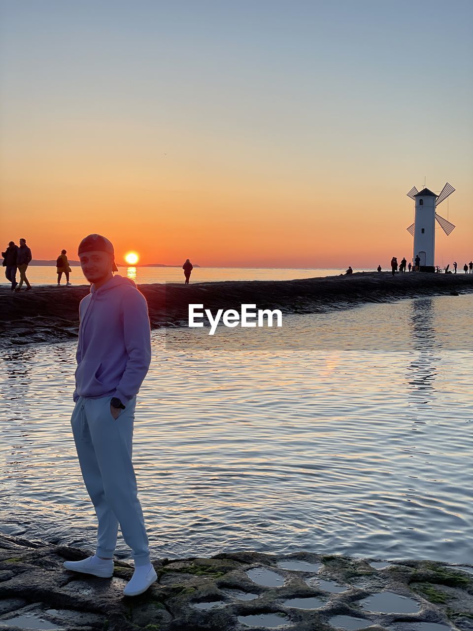 REAR VIEW OF MEN STANDING AT BEACH DURING SUNSET