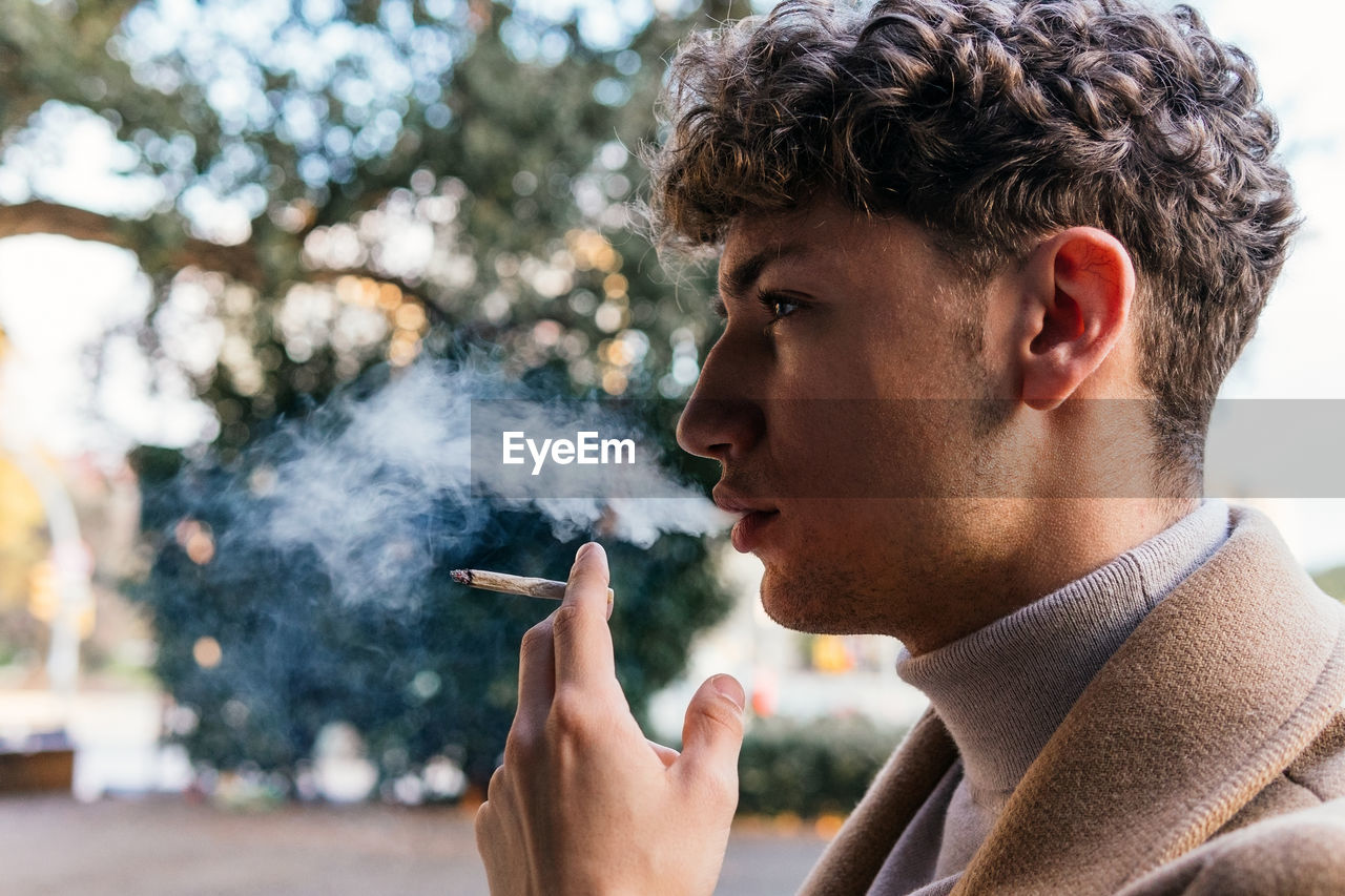 Side view of serious male in stylish outfit standing in street and smoking cigarette while exhaling fume and looking away