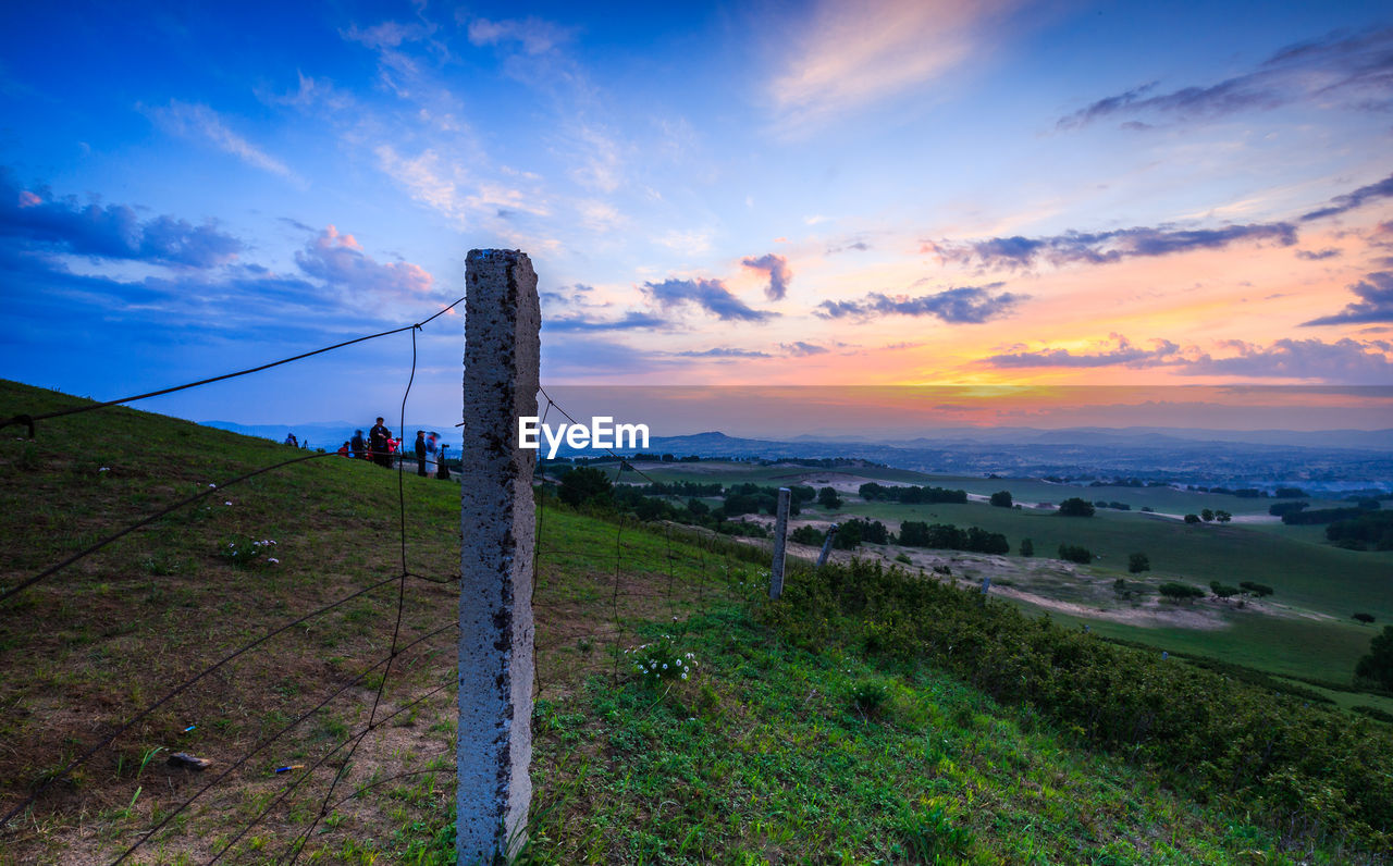 Railing on grassy hill against sky during sunset