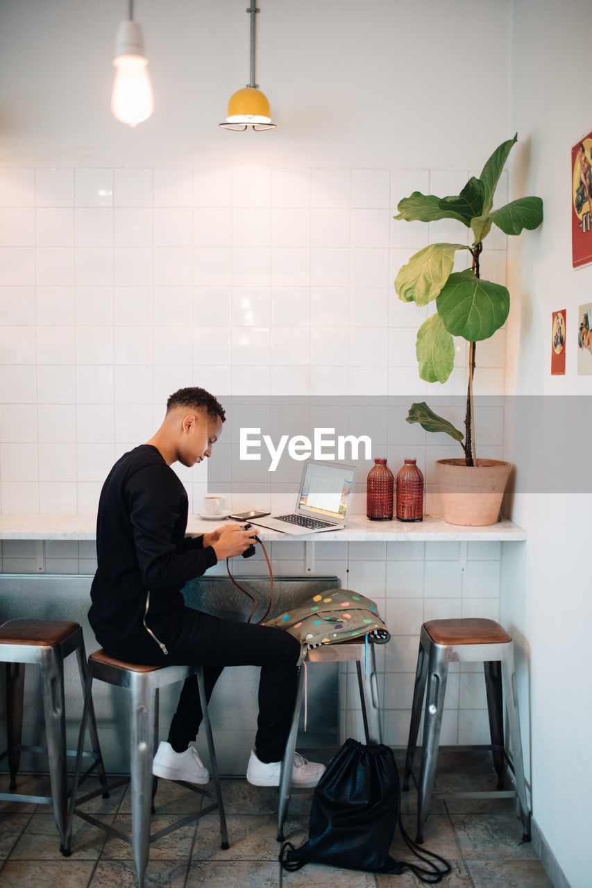 Full length of young male blogger sitting with camera and laptop at cafe