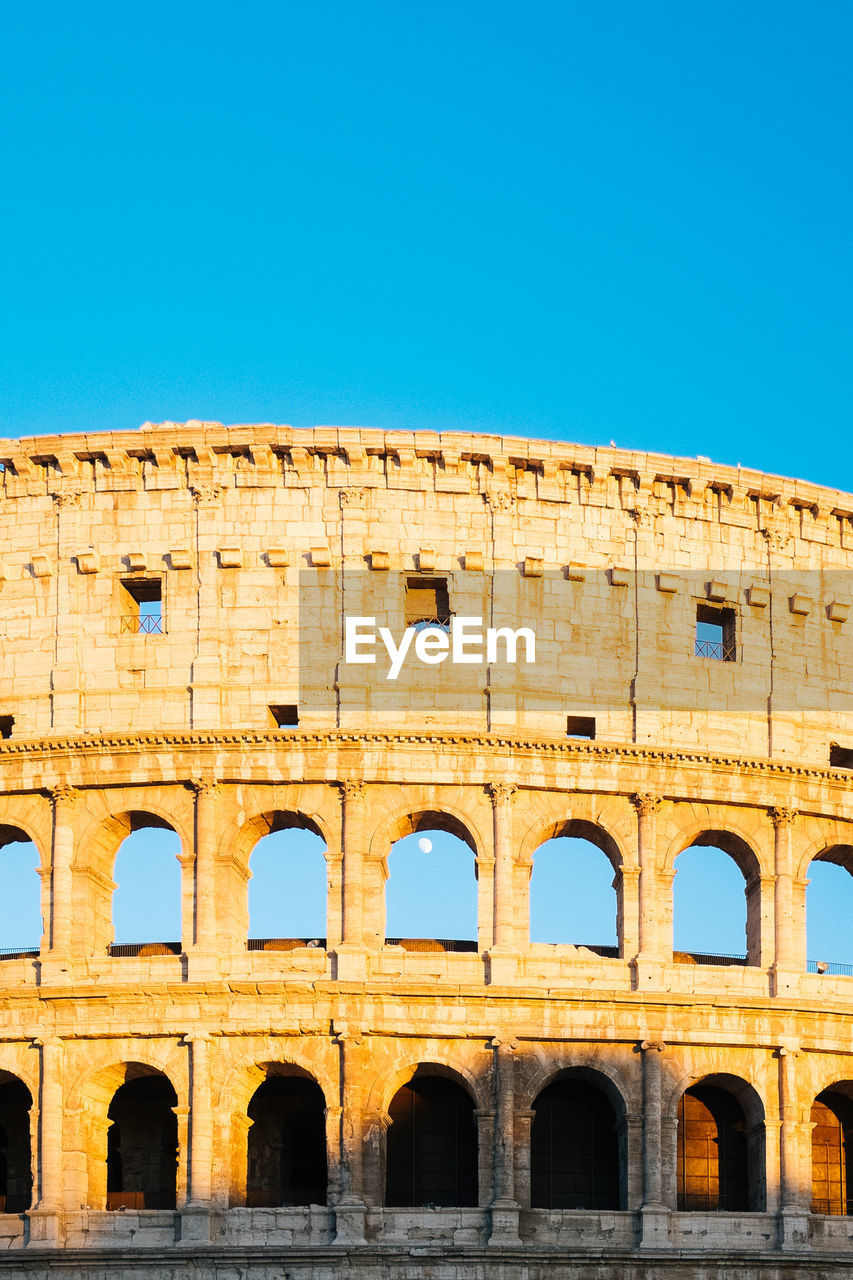 Low angle view of coliseum against clear blue sky on sunny day