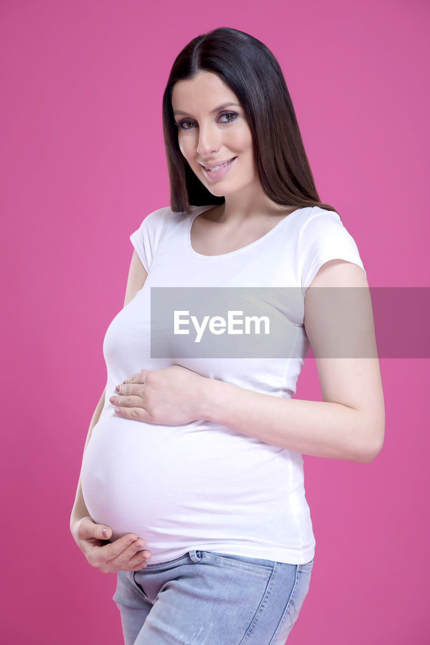 Portrait of smiling pregnant woman holding abdomen against pink background