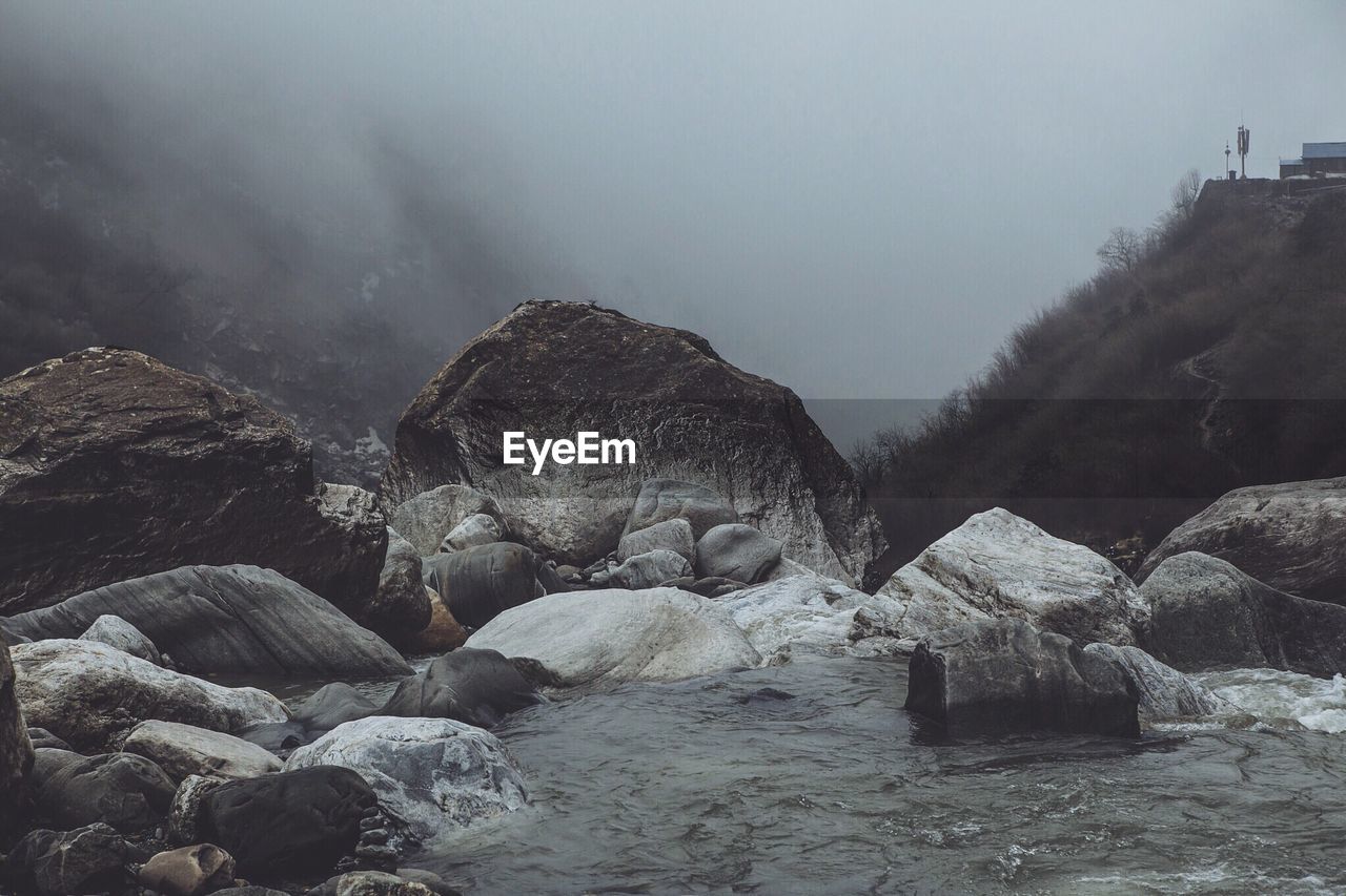 River flowing amidst rocks by mountain during foggy weather