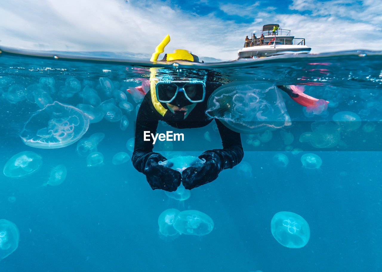 A woman in a black wetsuit is in the water with jellyfish. the jellyfish are floating around her