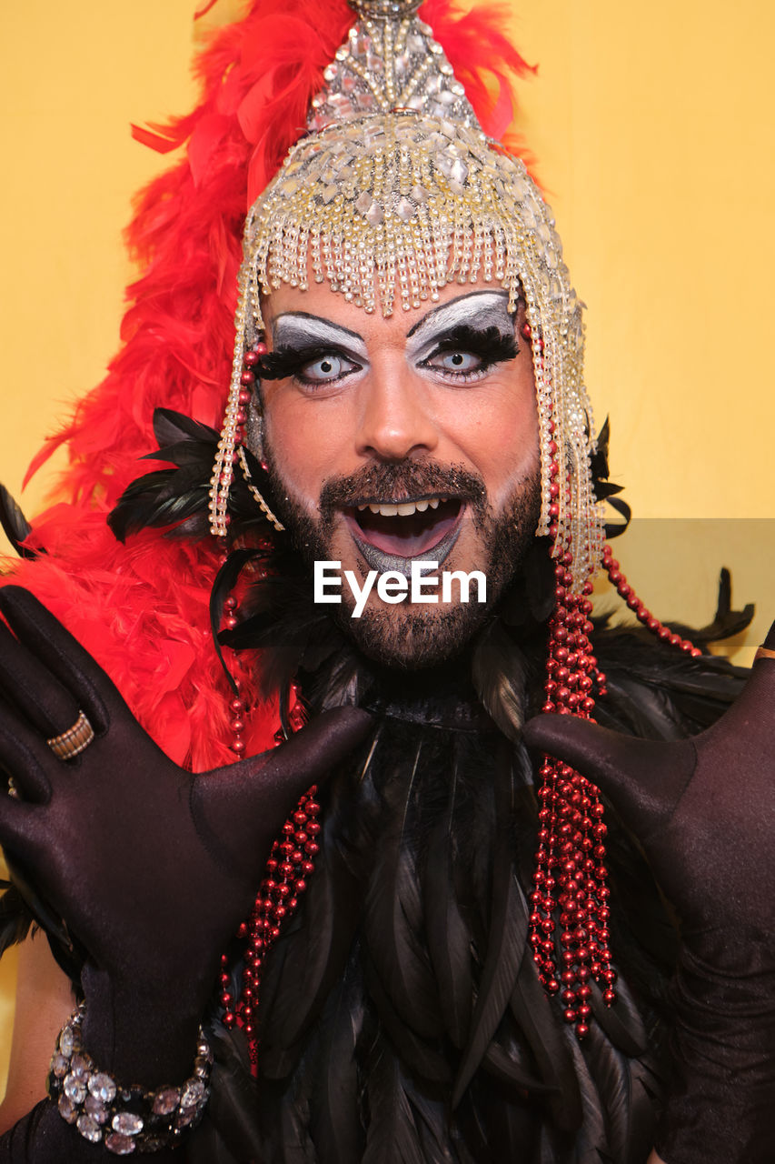 Extravagant bearded transgender male model with makeup wearing bodysuit and headwear with red and black feathers keeping hands on waist and looking at camera against yellow background