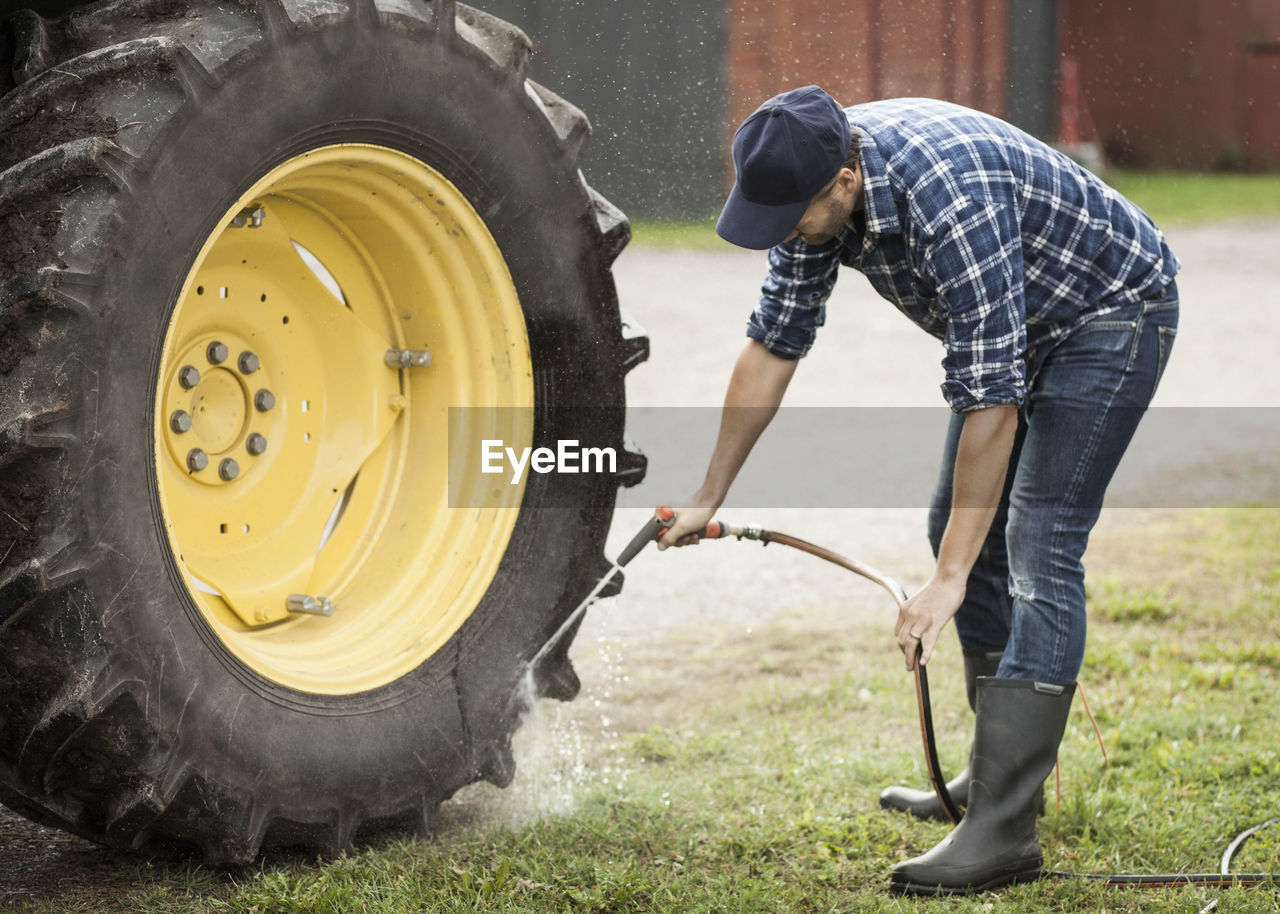 Full length of man washing tractor wheel with hose in farm