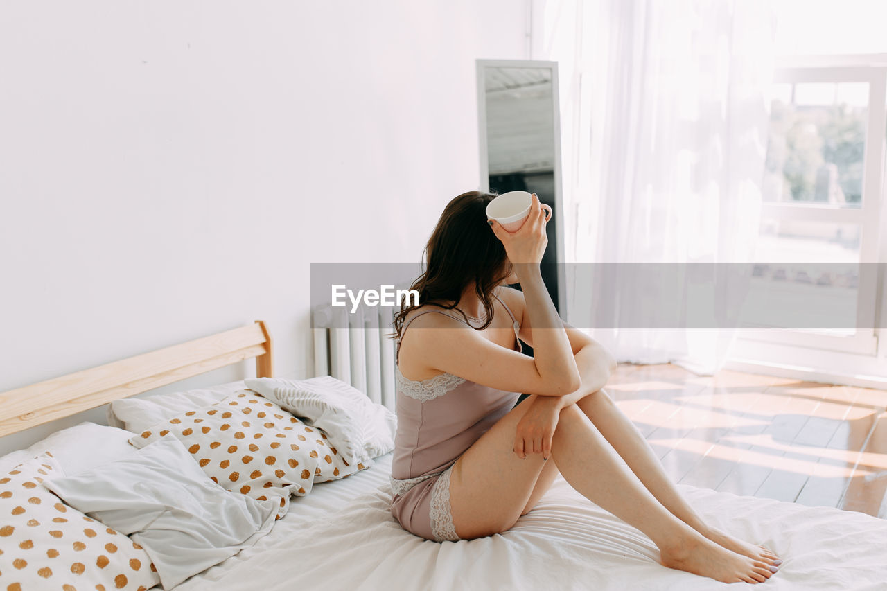 A happy girl in pajamas drinks coffee looks at the phone using technology while lying in bed at home