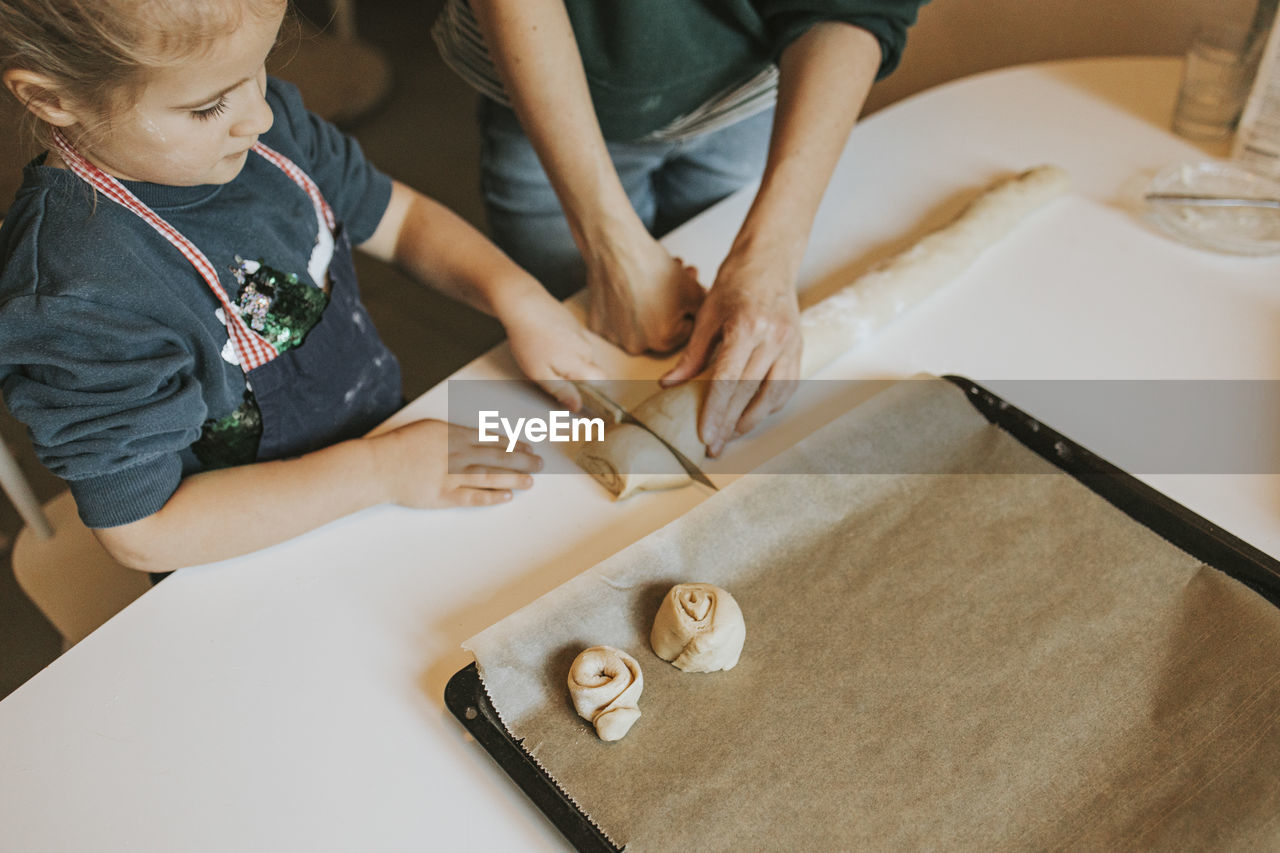 Mother holding cinnamon roll dough while daughter cutting with knife at table