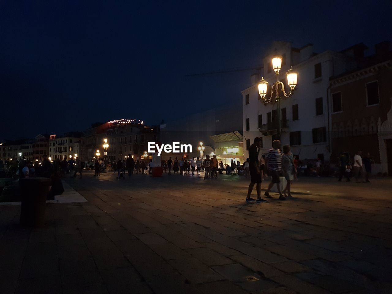 PEOPLE ON ILLUMINATED STREET AMIDST BUILDINGS AT NIGHT