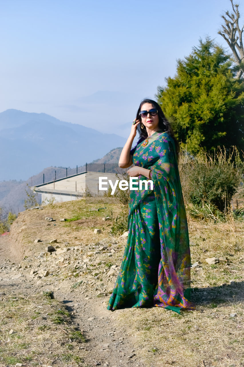 adult, full length, clothing, one person, fashion, women, dress, traditional clothing, nature, standing, portrait, young adult, green, blue, mountain, sky, land, photo shoot, front view, day, plant, outdoors, sari, looking at camera, landscape, smiling, female, tradition, looking, lifestyles, sunlight, leisure activity, environment, travel