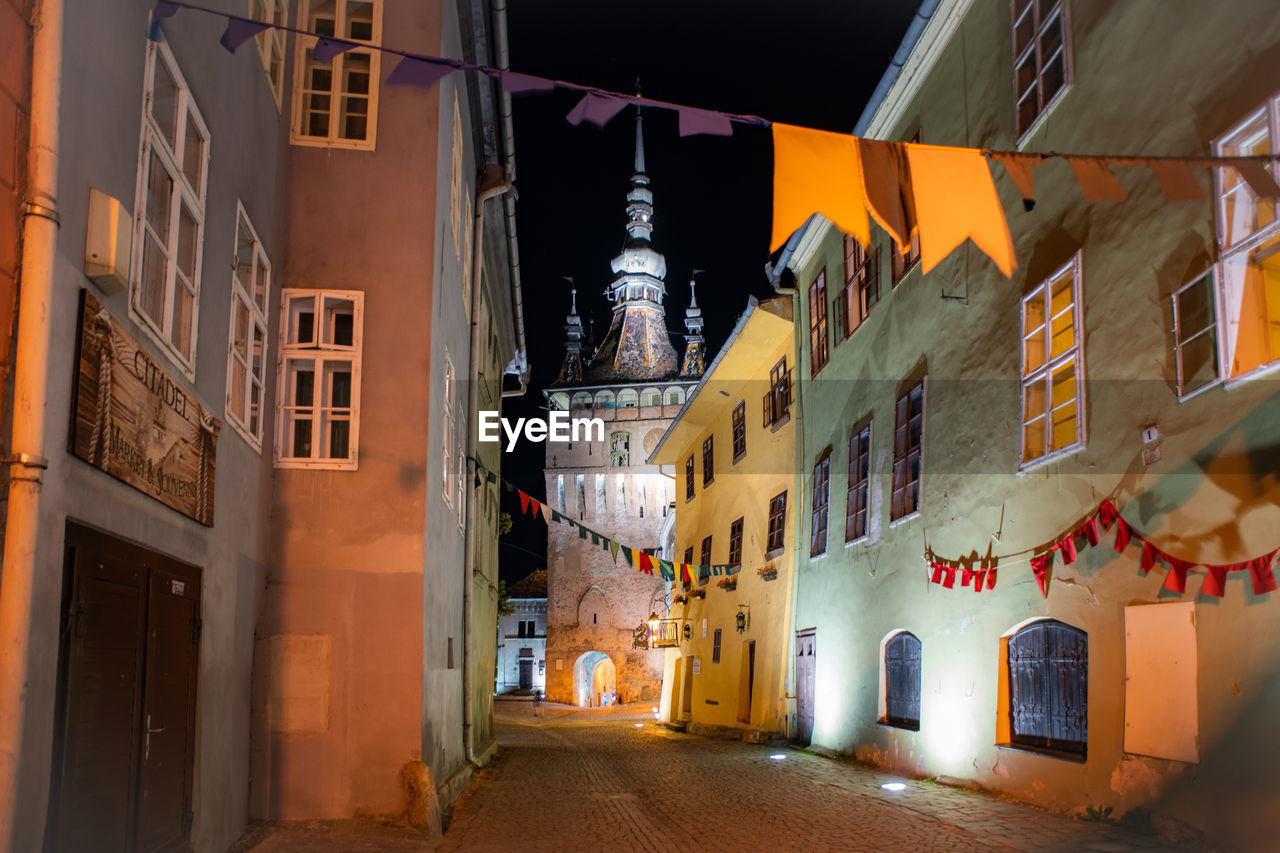 Narrow street amidst buildings in city at night. view of medieval clock tower in sighisoara, romania 